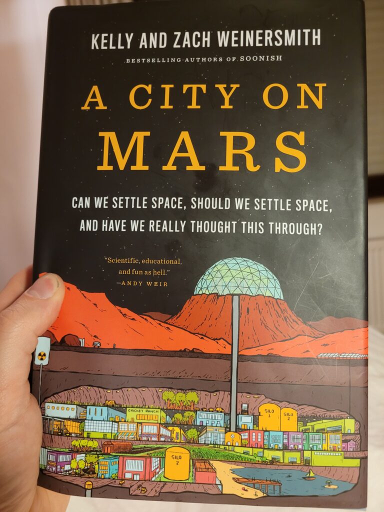 The cover of A City on Mars: Can we settle space, should we settle space, and have we really thought this through?, by Kelly&Zach Weinersmith. On the top half, in front of a starry background, the authors, title and subtitle. Below that, a martian landscape with a large, domed crater in the backgound and in the foreground a small exit with a vehicle and a human on the left and a bunch of solar arrays on the right. Below the surface, a cut-out view from an underground settlement with a central shaft going up the crater from the backgound and a smaller utility shaft going to a close-to-the-surface nuclear reactor. The settlement goes over multiple stories with small-town, medium-density vibes: agriculture on the top floor, below that an industrial floor including two silos and a cricket ranch, below that a living-quarters street with apartement-buildings, another layer of agriculture and a commercial floor, including an underground recreational lake and a Walmars (sic).