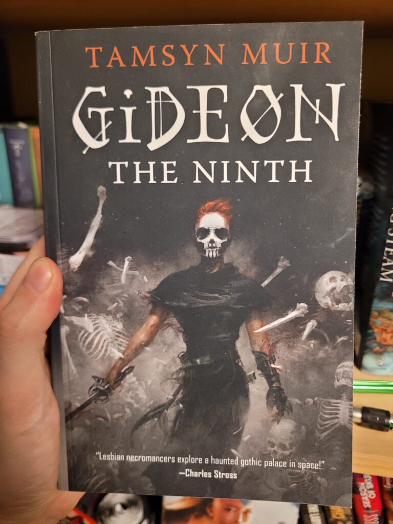 The cover of Gideon the Ninth, by Tamsyn Muir. It shows the eponymous desaster lesbian, a lean and muscular woman mostly clad in the black garb of the Ninth House but with uncovered arms, in her right hand a rapier and on her left a glove with steel claws. Her face is painted with a skull mask beneath short red hair in a crew cut. Across her back is strapped (heh.) a sword, with the hilt extending over her left shoulder. In the background a whole lot of skeletons are in the process of shattering explosively.