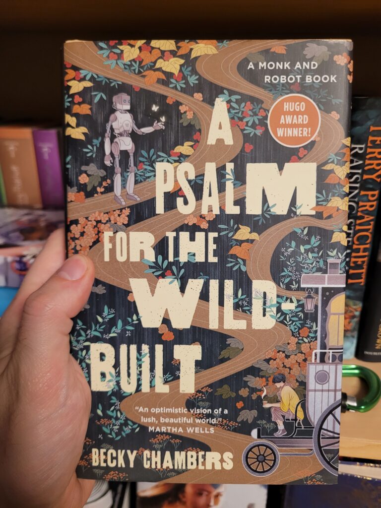 The cover of A Psalm for the Wild-Built, by Becky Chambers. In shows the title in varied-width-letters over a semi-stylized forest crossed winding roads and pathways, with Mosscap standing on a path playing with butterflies and Sibling Dex sitting on their wagon with a cup of tea in their hands.