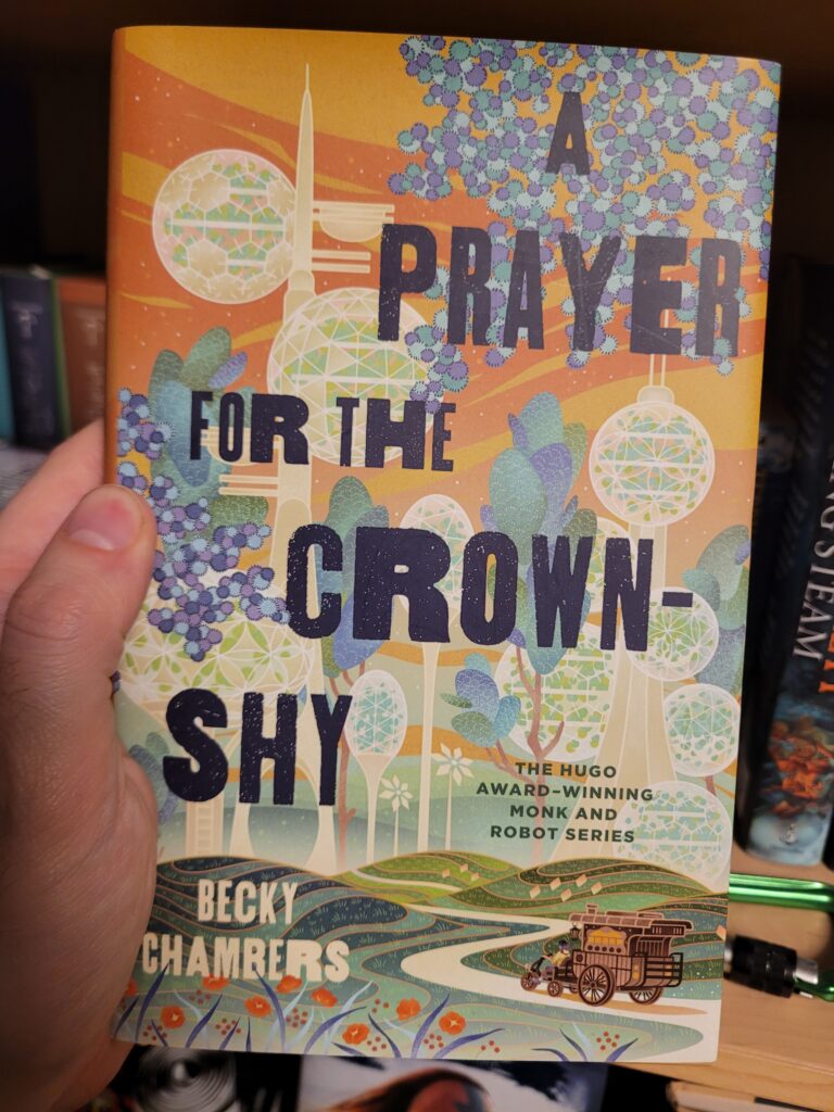 The cover of A Prayer for the Crown-Shy, by Becky Chambers. It shows the title in width-varied letters in front of a picture, in the background of which a stylized view of a city of habitat-spheres on poles, growing amid the trees of a forest, in the foreground a road winding through some hills, on it the tea-wagon of Sibling Dex.