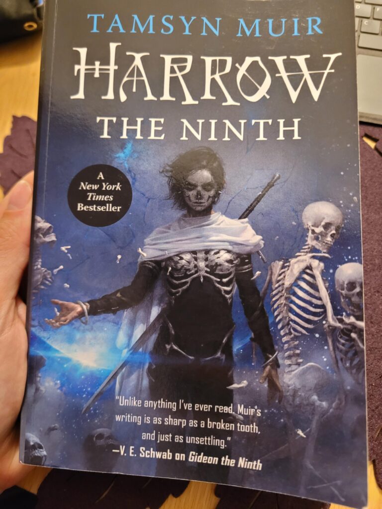 The cover of Harrow the Ninth, by Tamsyn Muir. In the background the gray of a dead planet in front of stars, with blue veins across it and a blue star barely flaring across its lower edge. In front of that the protagonist, Lady Harrowhark, The Revered Daughter, a young women with short black hair and a skull painted on her face, clad in black garments, bones forming a ribcage over her chest, and a white coat slung over her shoulders. Across her back she carries a large, two-handed sword. Around her are rather less than more complete skeletons, skulls and bones pieces, floating around her and her outstretched hand.