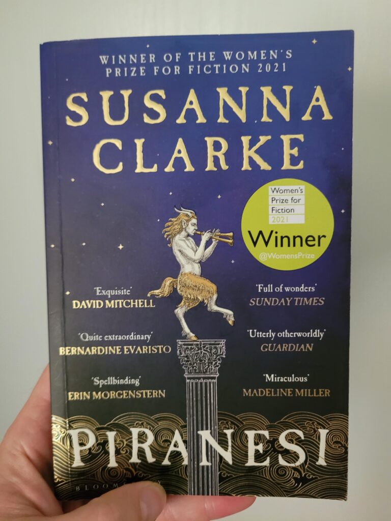 The cover of Piranesi, by Susanna Clarke. In front of a dark blue night sky with golden stars stands a column, on which a (a statue of a?) faun stands upright with one hoof, the other raised in the air. The faun has pale skin and blond hair on its head and its lower body and legs, and it's playing a double-ended flute. Along the bottom of the cover are the stylized swirls of a roaring ocean. The name of the author is printed on the top, the name of the book on the bottom.