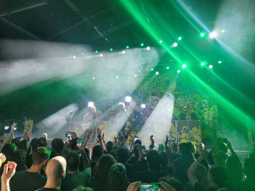 Ein Photo des WiZo-Konzerts: im Vordergrund die Menge, auf der Bühne die Band mit Lampen auf dem Rücken, um das Licht zu bringen.